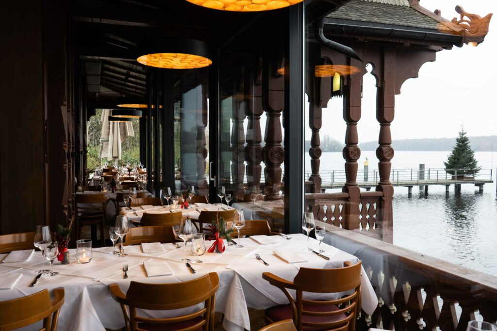 Überdachte Restaurantterrasse mit weiß gedeckten Tischen, warmen Deckenleuchten und Blick auf den See.