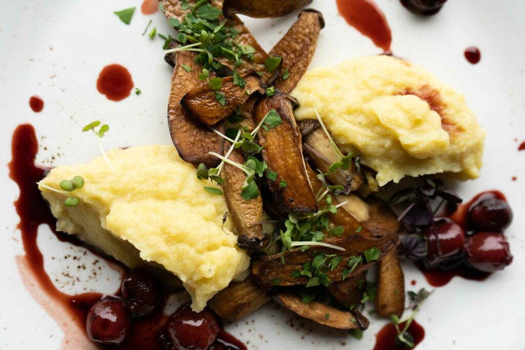 Gericht mit Kartoffelpüree, gebratenen Pilzen, Kräutern und dunkler Fruchtsauce.