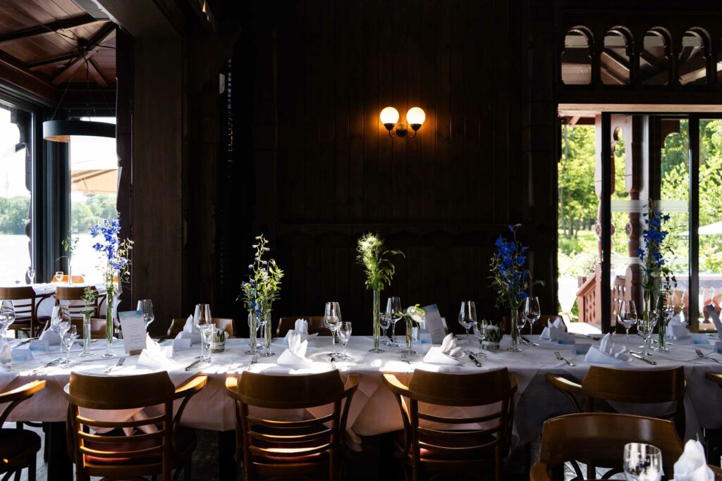 Festlich gedeckter Restaurantbereich mit langen Tafeln, Blumenarrangements und Blick ins Grüne.