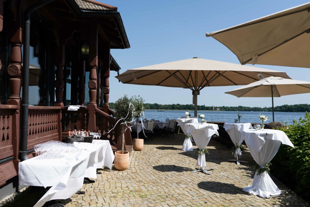 Außenterrasse mit weiß gedeckten Tischen, Sonnenschirmen und direktem Blick aufs Wasser.