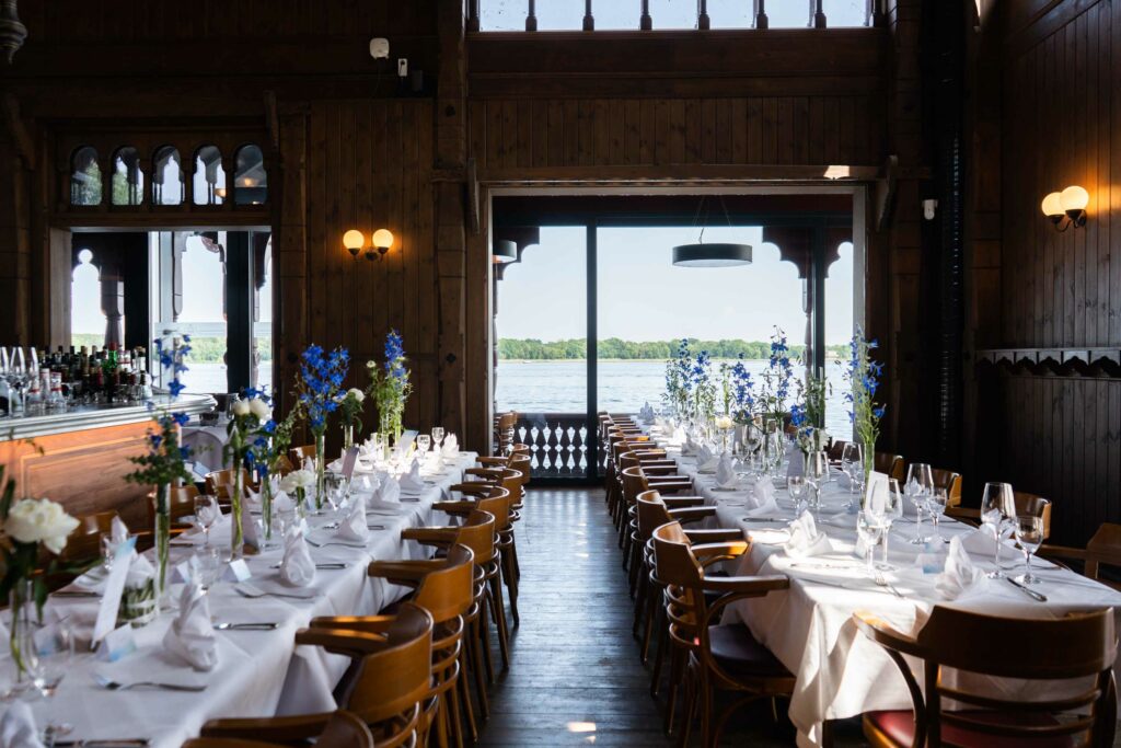 Langer festlich gedeckter Tisch im Restaurant mit Blumen, Gläsern und Blick auf den See.