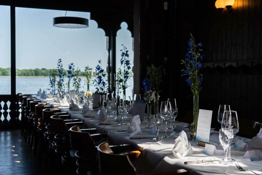 Restaurantinnenraum mit weiß eingedeckten Tischen, blauen Blumen und großen Fensterfronten.