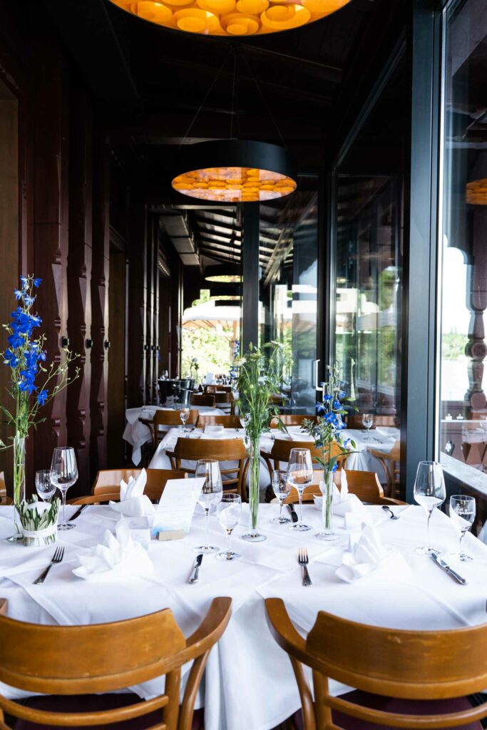 Restaurantinnenraum mit weiß eingedeckten Tischen, gefalteten Servietten, Weingläsern und blauen Blumen vor großen Fensterfronten.