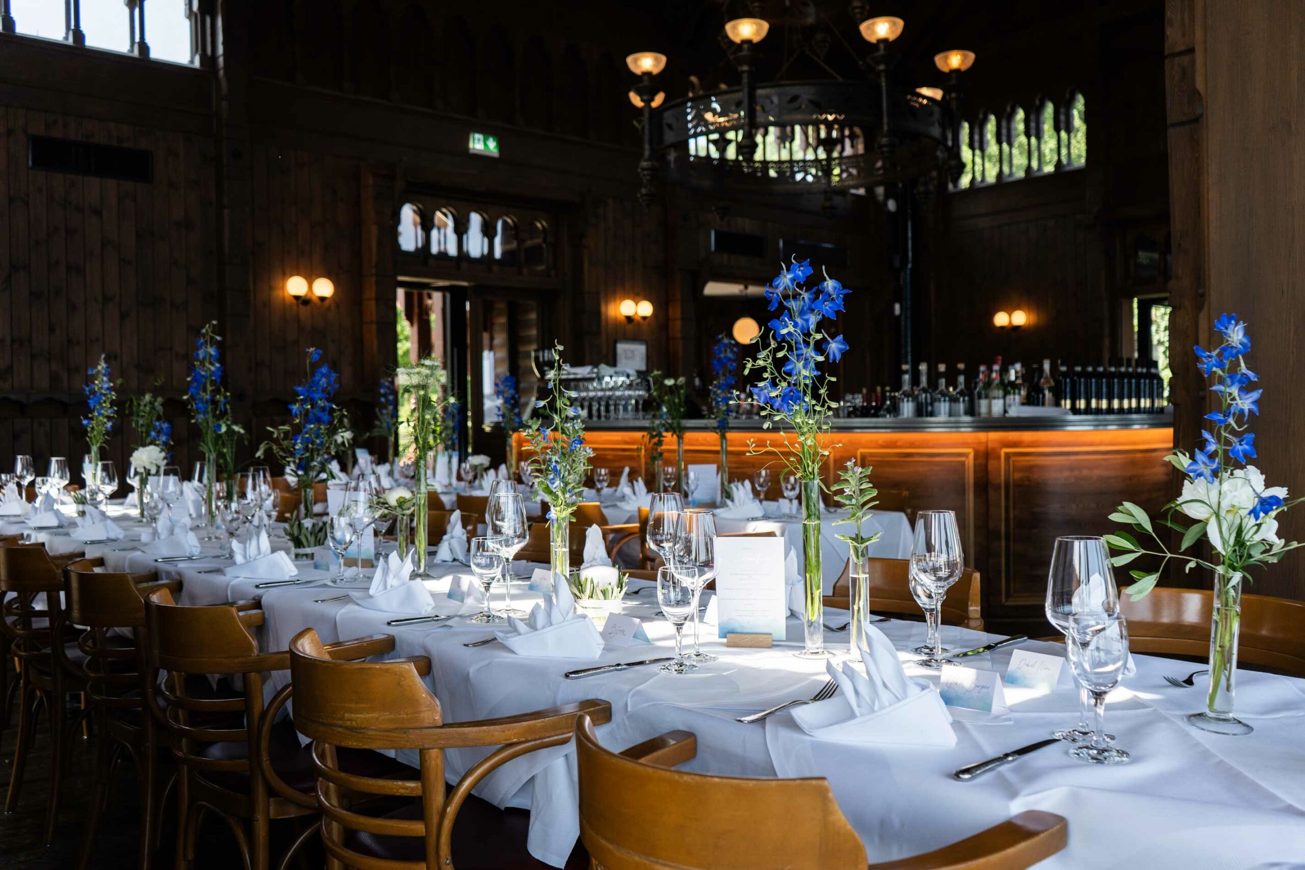 Festlich gedeckter Restaurantsaal mit weißen Tischdecken, Weingläsern, blauen Blumen und großer Holzbar im Hintergrund.