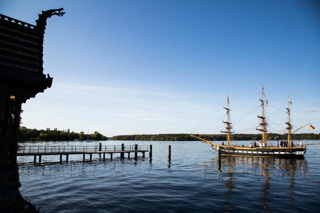 Seeblick mit Steg, Geländer und Segelbooten bei klarem Himmel.