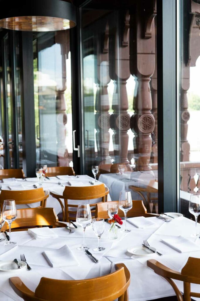 Restaurantinnenraum mit weiß eingedeckten Tischen, Gläsern und Holzmöbeln vor großen Fenstern mit Terrassenblick.