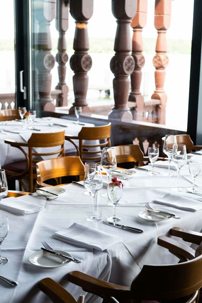 Innenraum eines Restaurants mit weiß eingedeckten Tischen, Weingläsern und Holzmöbeln vor großen Fenstern mit Blick auf eine Terrasse.