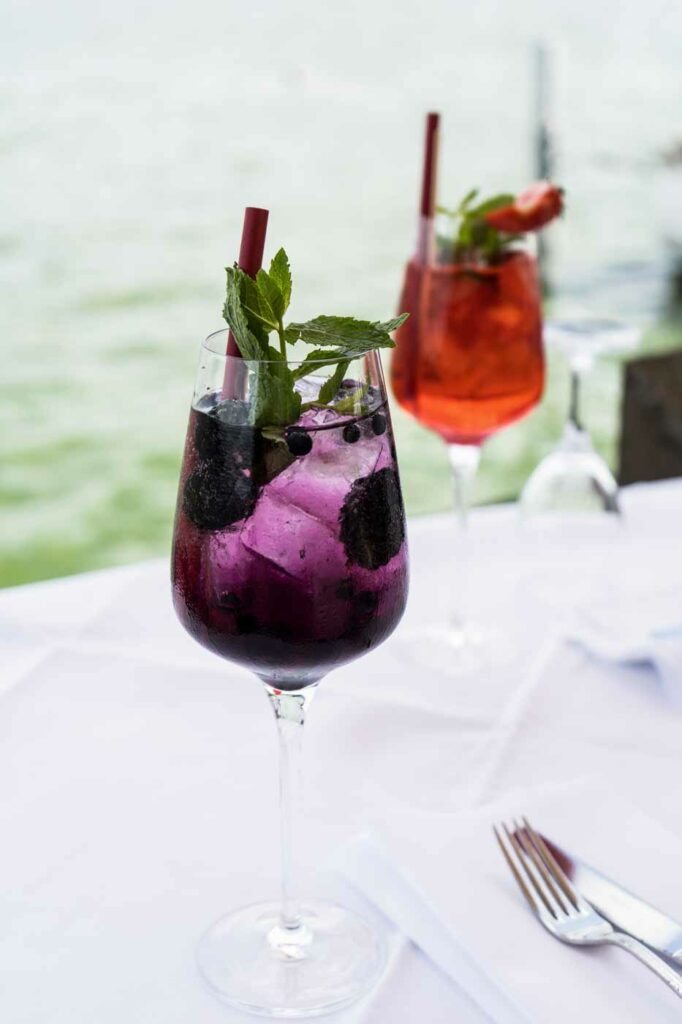 Zwei farbige Cocktails in Gläsern mit frischer Garnitur auf einem weiß gedeckten Tisch.