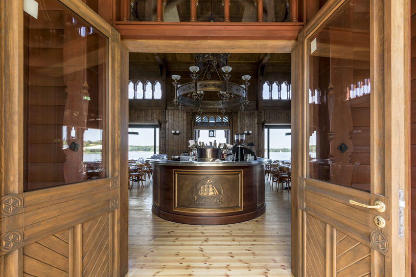 Restaurantinnenraum mit großer Holzbar, Holzwänden, gedeckten Tischen und warmem Licht.