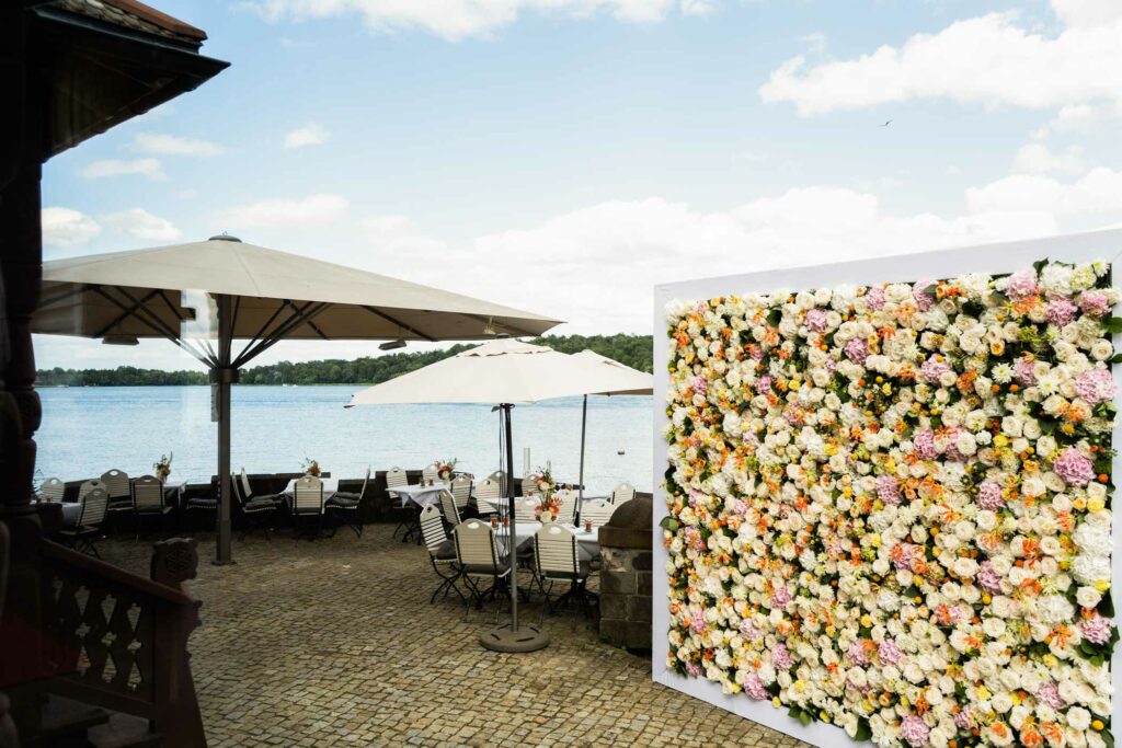 Seeterrasse mit gedeckten Stehtischen, Blumenwand und Sonnenschirmen.
