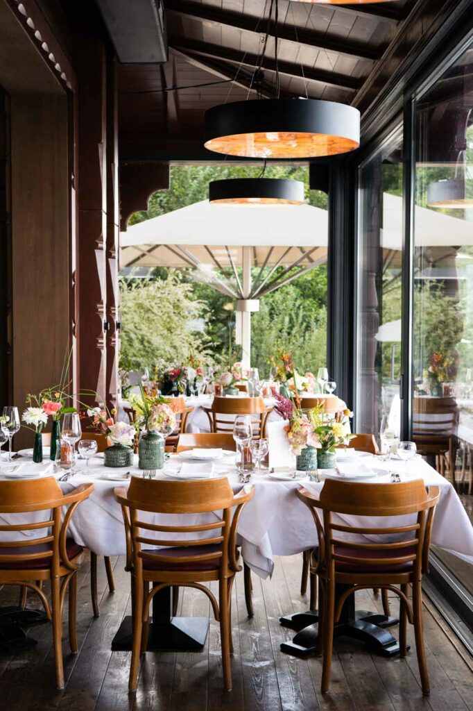 Festlich gedeckte Restauranttische mit Blumenarrangements, Holzmöbeln und großen Fensterfronten mit Blick auf die Terrasse.