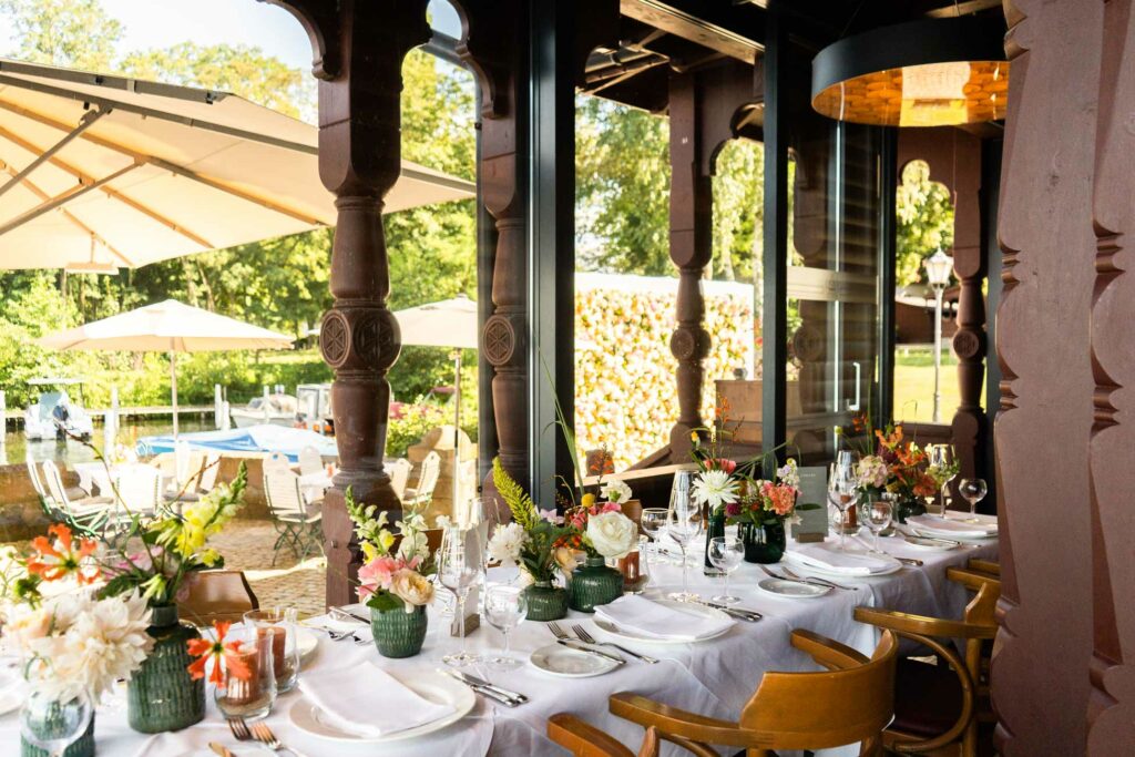 Festlich gedeckter Restaurantbereich mit Blumenarrangements, großen Fenstern und Blick auf die Terrasse.