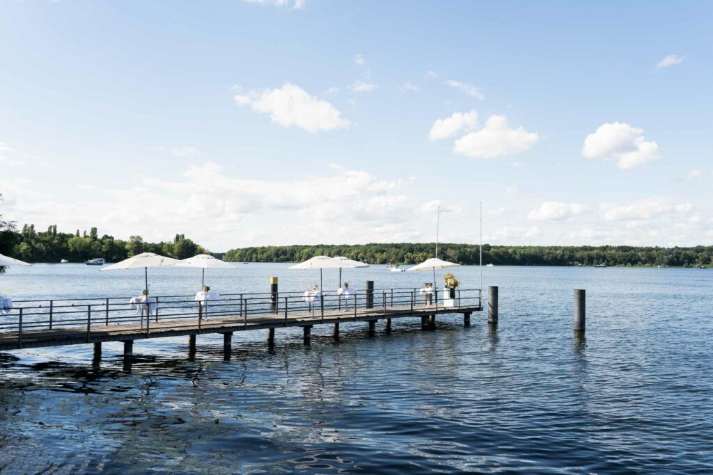 Seeblick mit langem Steg, Geländer und ruhigem Wasser unter blauem Himmel.
