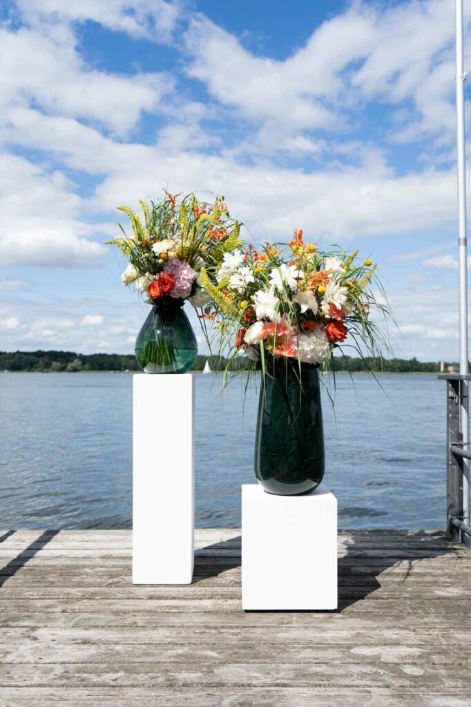Zwei große Blumenarrangements in dunklen Glasvasen auf weißen Säulen vor weitem Seeblick und blauem Himmel.