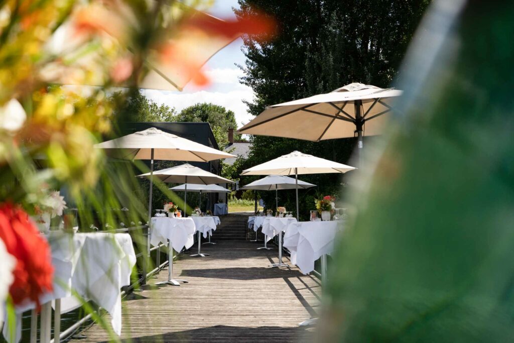 Sonnige Terrasse mit Sonnenschirmen, Pflanzen und Blick auf das Restaurant am See.