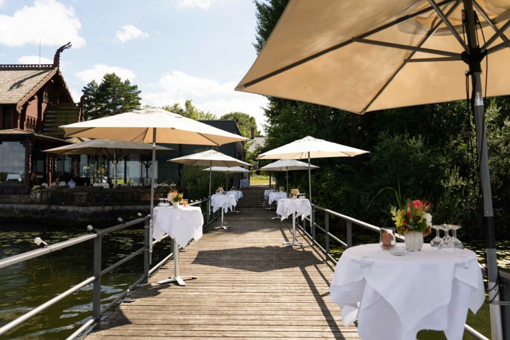 Terrasse am See mit Sonnenschirmen, weiß gedeckten Tischen und Blick ins Grüne.