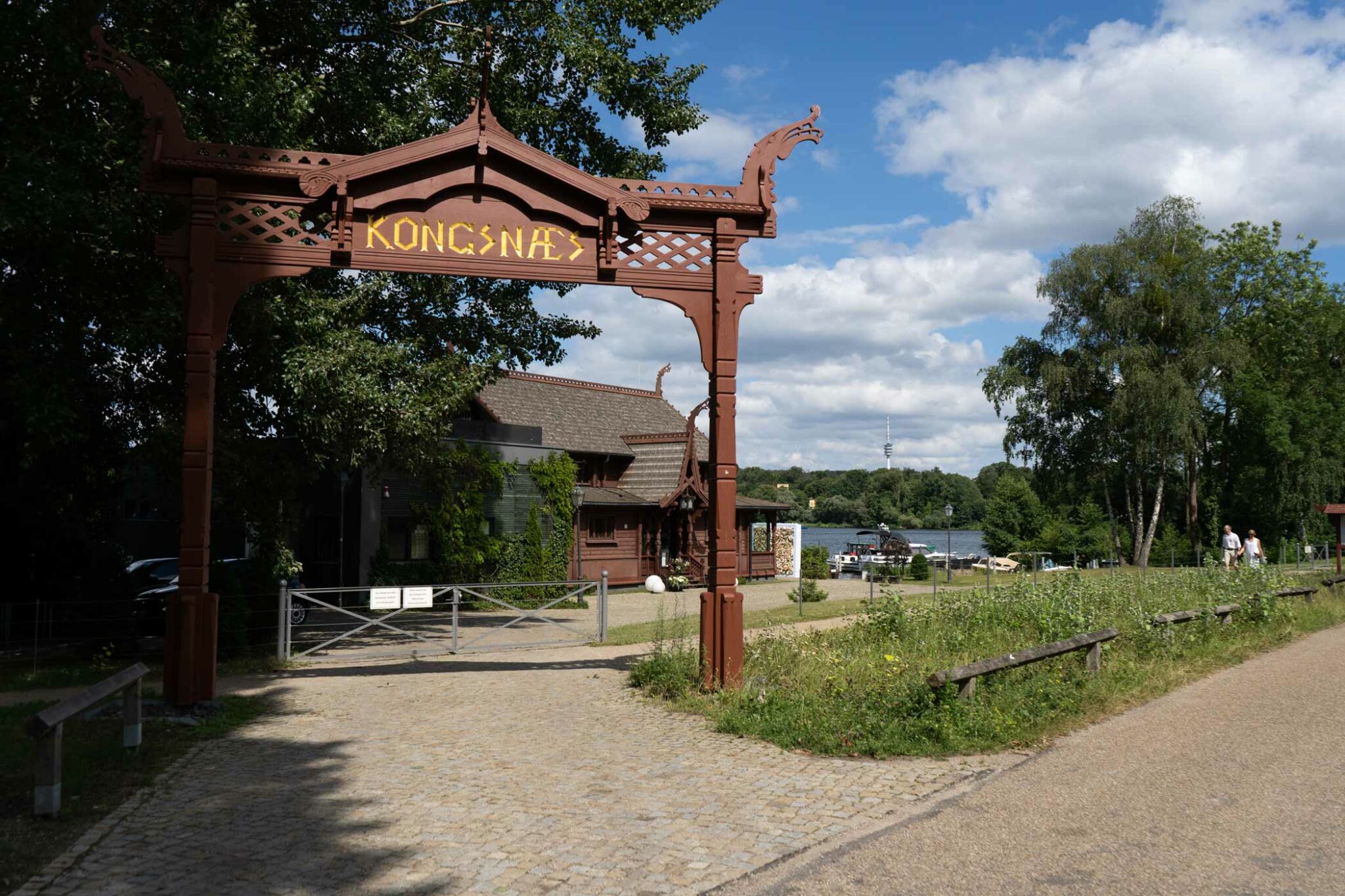 Eingangstor mit dekorativer Beschriftung, Weg und Grünfläche vor einem Gebäude am See.