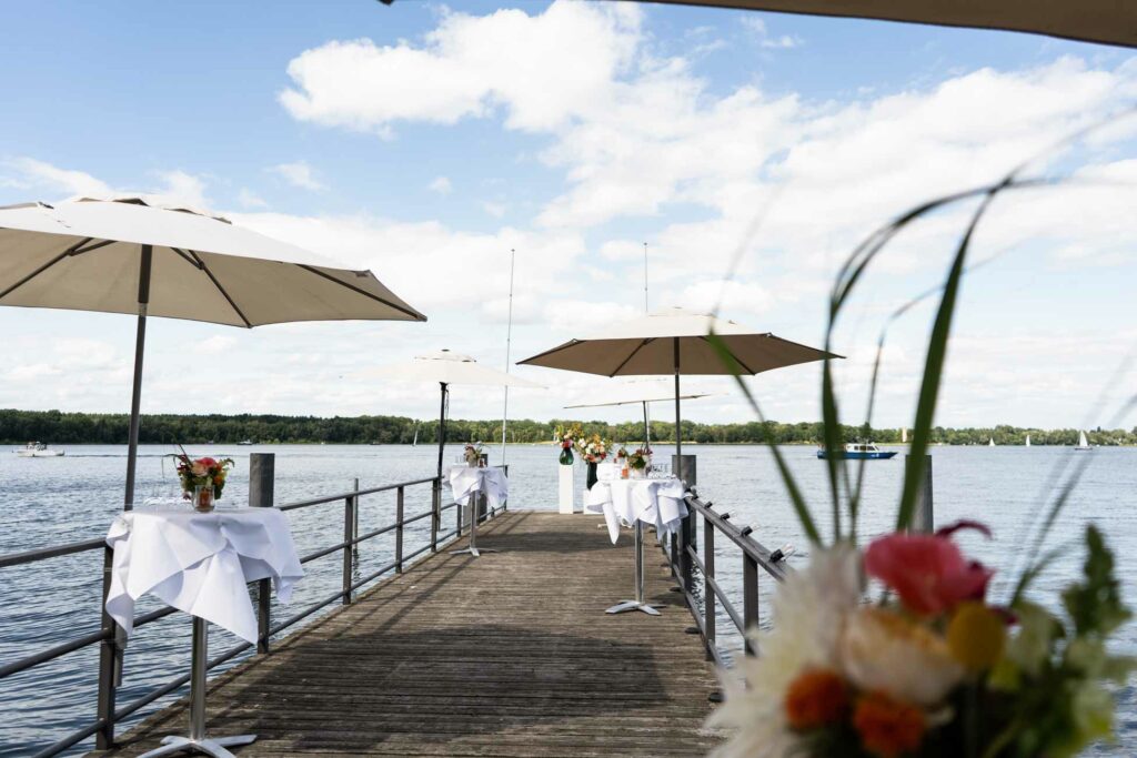 Seeterrasse mit Sonnenschirm, Blumenschmuck und gedeckten Tischen am Wasser.